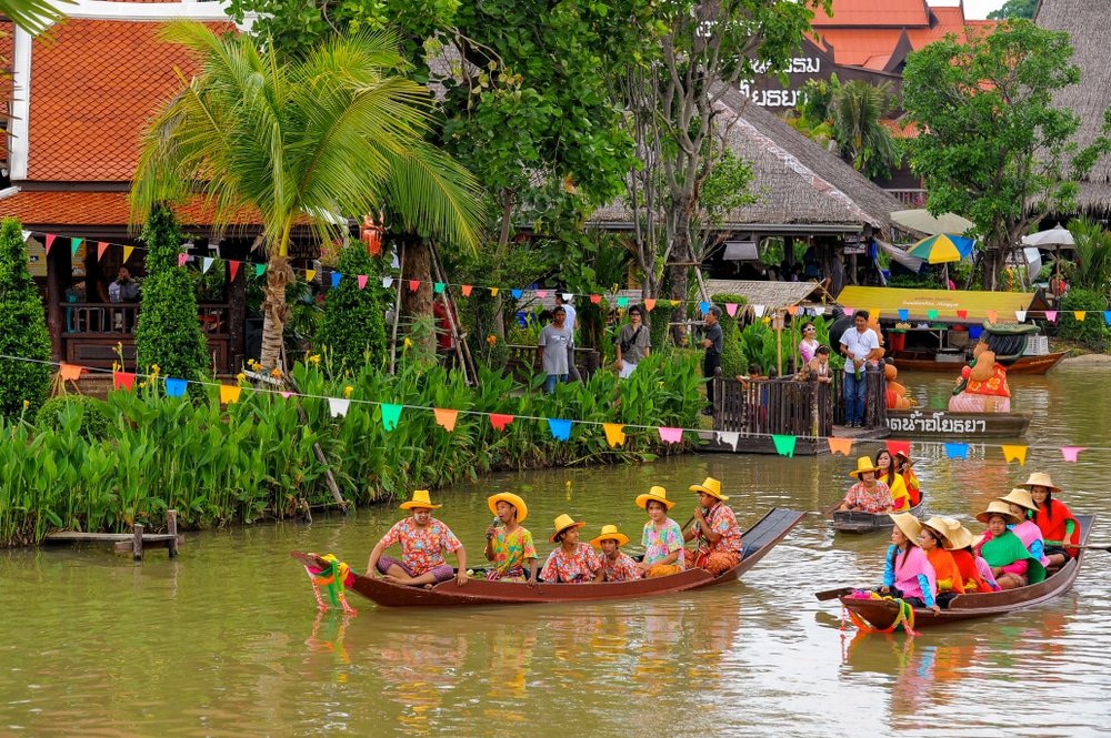 ผู้คนในชุดสีสันสดใสและหมวกฟางพายเรือไม้ไปตามคลอง ท่ามกลางธรรมชาติสีเขียวและอาคารที่ตกแต่งอย่างสวยงาม โดยมีผู้คนที่มายืนชมอยู่ริมน้ำ ณ สถานที่ท่องเที่ยวอันมีเสน่ห์ในอยุธยา.