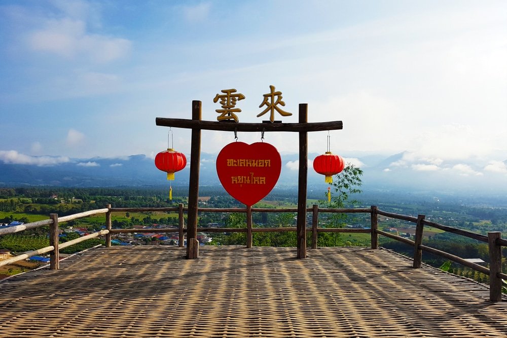 แพลตฟอร์มชมวิวไม้พร้อมโคมไฟสีแดงและป้ายรูปหัวใจ มองเห็นทิวทัศน์ที่สวยงามของภูเขาและท้องฟ้าสีคราม—เหมาะสำหรับการถ่ายภาพที่สถานที่ท่องเที่ยวอยุธยานี้.