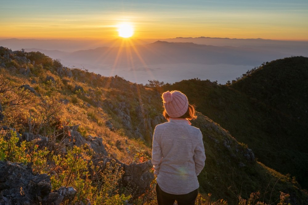 บุคคลสวมหมวกสีชมพูและเสื้อแจ็คเก็ตบางยืนอยู่บนเนินเขาที่ปกคลุมด้วยหญ้า กำลังชมแสงอาทิตย์ยามเช้าที่ส่องประกายเหนือเนินเขาไกล—ช่วงเวลาที่สมบูรณ์แบบ ณ สถานที่ท่องเที่ยวเชียงใหม่ที่มีชื่อเสียงในด้านความงดงามอันเงียบสงบ.