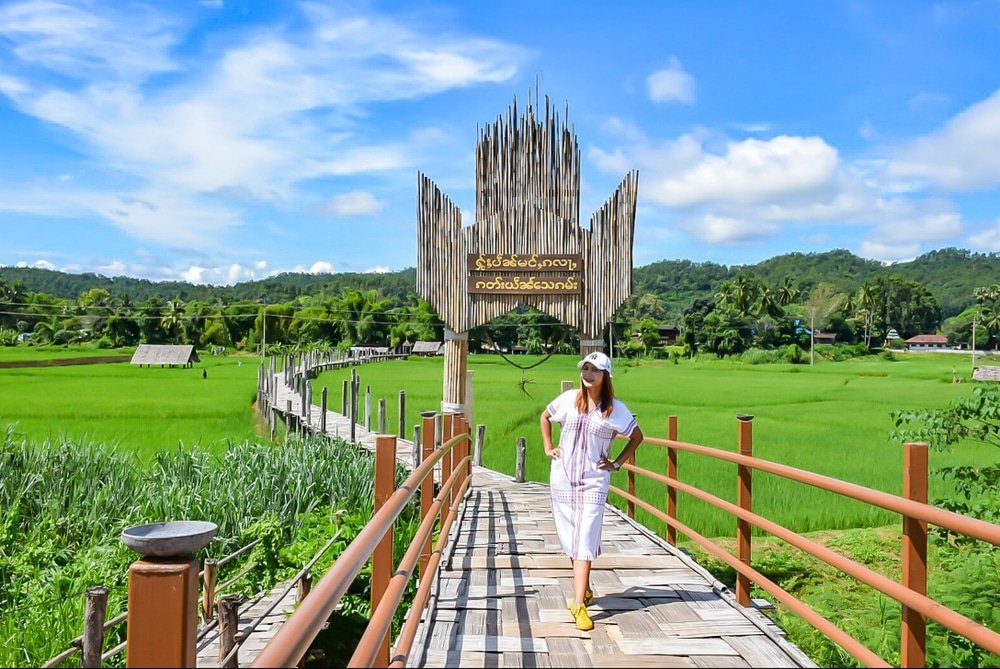 ผู้หญิงในชุดสีขาว ยืนอยู่บนสะพานไม้เหนือทุ่งนาข้าวสีเขียว โดยมีป้ายขนาดใหญ่รูปมืออยู่ด้านหลัง ภายใต้ท้องฟ้าสีคราม ณ สถานที่ท่องเที่ยวอยุธยา.