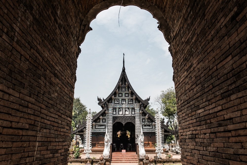 วัดแบบดั้งเดิมที่มีรายละเอียดประณีตถูกมองผ่านซุ้มประตูอิฐ ขนาบข้างด้วยรูปปั้นสีขาวและล้อมรอบด้วยต้นไม้ใต้ท้องฟ้าที่มีเมฆบางส่วน—สถานที่ที่ต้องไปเยือนสำหรับใครก็ตามที่กำลังมองหา ที่เที่ยวเชียงใหม่.