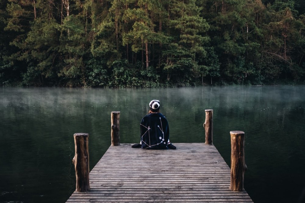 บุคคลหนึ่งสวมเสื้อคลุมมีฮู้ดนั่งขัดสมาธิอยู่ที่ปลายท่าเรือไม้ หันหน้าไปทางทะเลสาบที่มีหมอกปกคลุมและต้นไม้เขียวชอุ่มหนาแน่น—สร้างบรรยากาศที่เงียบสงบไม่แพ้สถานที่ท่องเที่ยวอยุธยา.