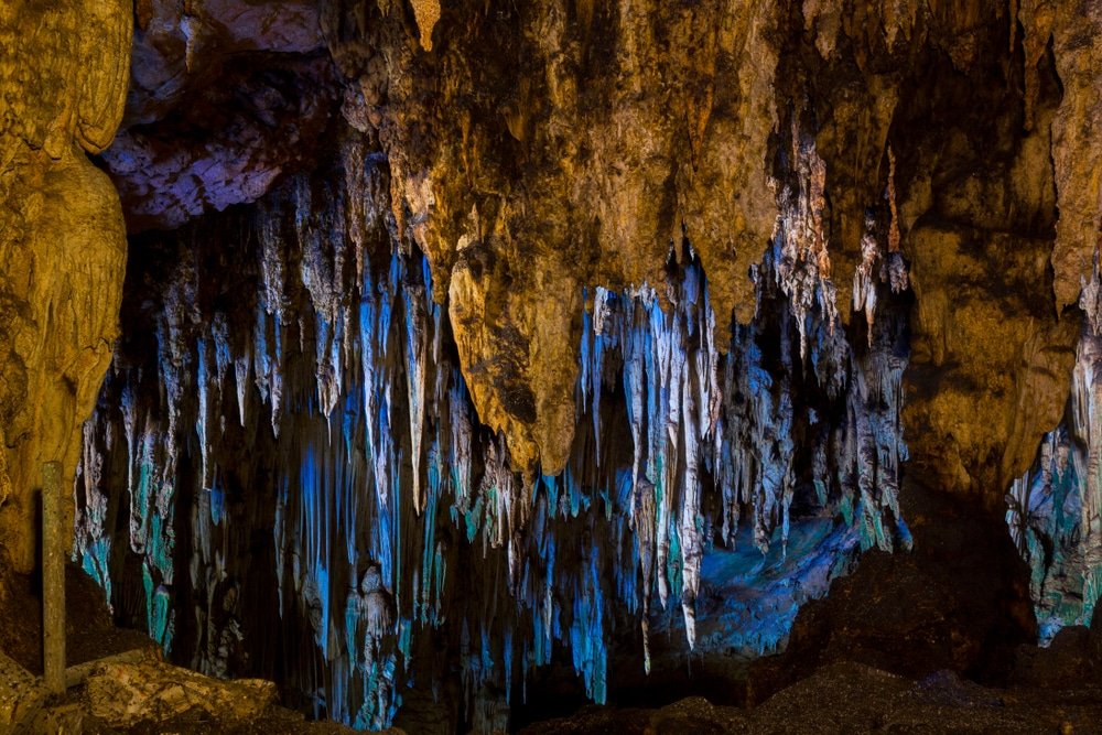 หินงอกห้อยลงมาจากเพดานของถ้ำ ส่องสว่างด้วยแสงสีฟ้าและสีเหลือง มีหินรูปร่างต่างๆ บนพื้นและผนัง—สถานที่ที่น่าหลงใหลเหมาะสำหรับผู้ที่กำลังมองหาสถานที่ท่องเที่ยวในอยุธยา.