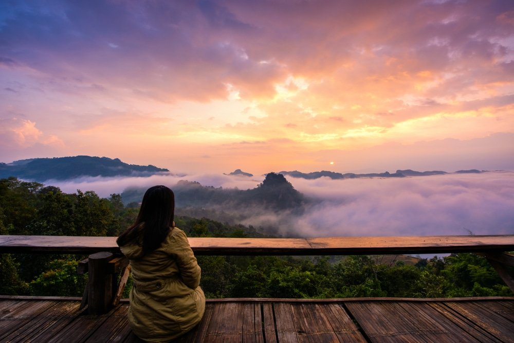 บุคคลในเสื้อแจ็กเก็ตนั่งอยู่บนแพลตฟอร์มไม้ มองดูทิวทัศน์ภูเขาที่ปกคลุมด้วยหมอกและท้องฟ้าที่เต็มไปด้วยสีสันของพระอาทิตย์ขึ้น ซึ่งชวนให้นึกถึงความงดงามอันเงียบสงบที่พบได้ที่ ที่เที่ยวอยุธยา.