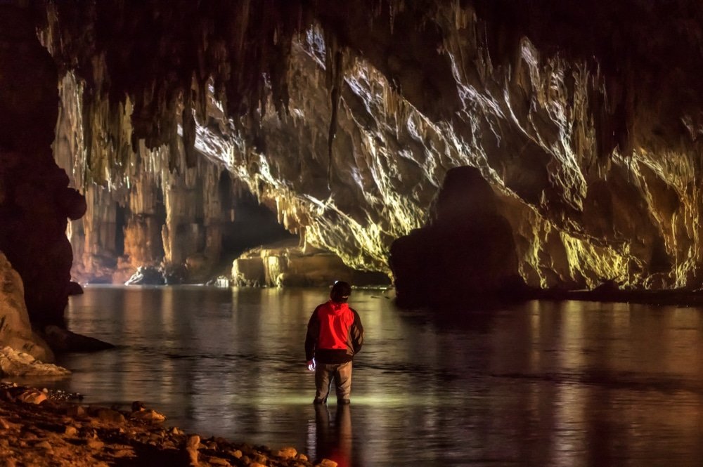 บุคคลที่สวมเสื้อแจ็คเก็ตสีแดงยืนอยู่ในน้ำตื้น หันหน้าไปทางภายในถ้ำขนาดใหญ่ที่ส่องสว่างด้วยแสงสว่างจากภายใน พร้อมด้วยหินรูปร่างแปลกตา—การผจญภัยที่ชวนให้นึกถึงการสำรวจมุมลับ ๆ ขณะเที่ยวอยุธยา.