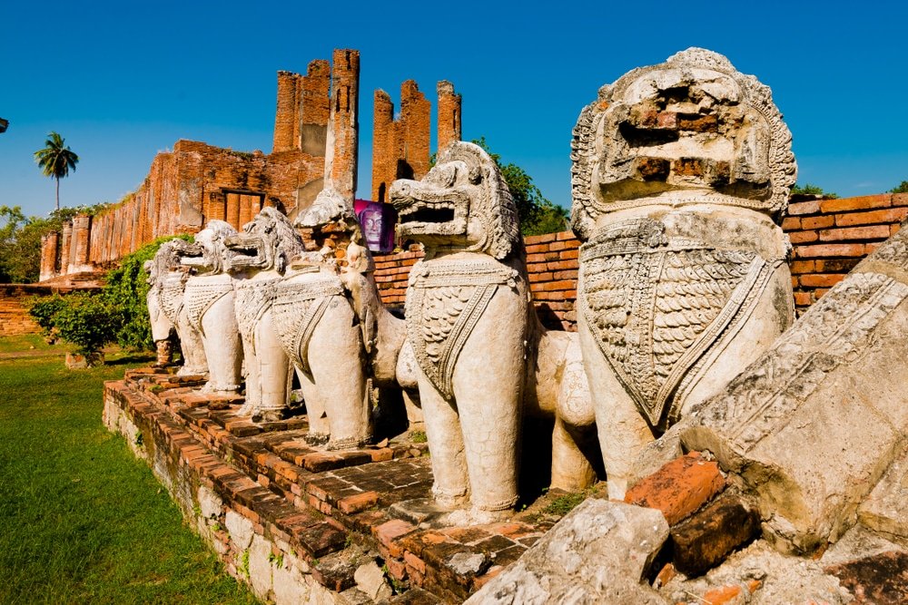 รูปปั้นสิงโตหินโบราณเฝ้าซากอิฐใต้ท้องฟ้าสีฟ้าใส ณ วัดประวัติศาสตร์แห่งนี้ สถานที่ท่องเที่ยวอยุธยาที่ต้องไปเยือน สำหรับผู้ที่ต้องการสำรวจความมหัศจรรย์ของการเที่ยวอยุธยา.