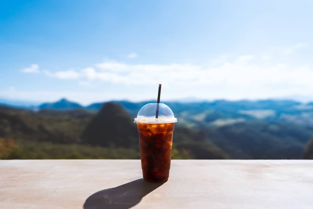 แก้วพลาสติกใส่น้ำแข็งกาแฟพร้อมหลอดวางอยู่บนพื้นผิวเรียบ มีภูเขาและท้องฟ้าแจ่มใสเป็นฉากหลัง—เหมาะสำหรับการผ่อนคลายหลังจากวันที่สำรวจอยุธยาที่เที่ยว.