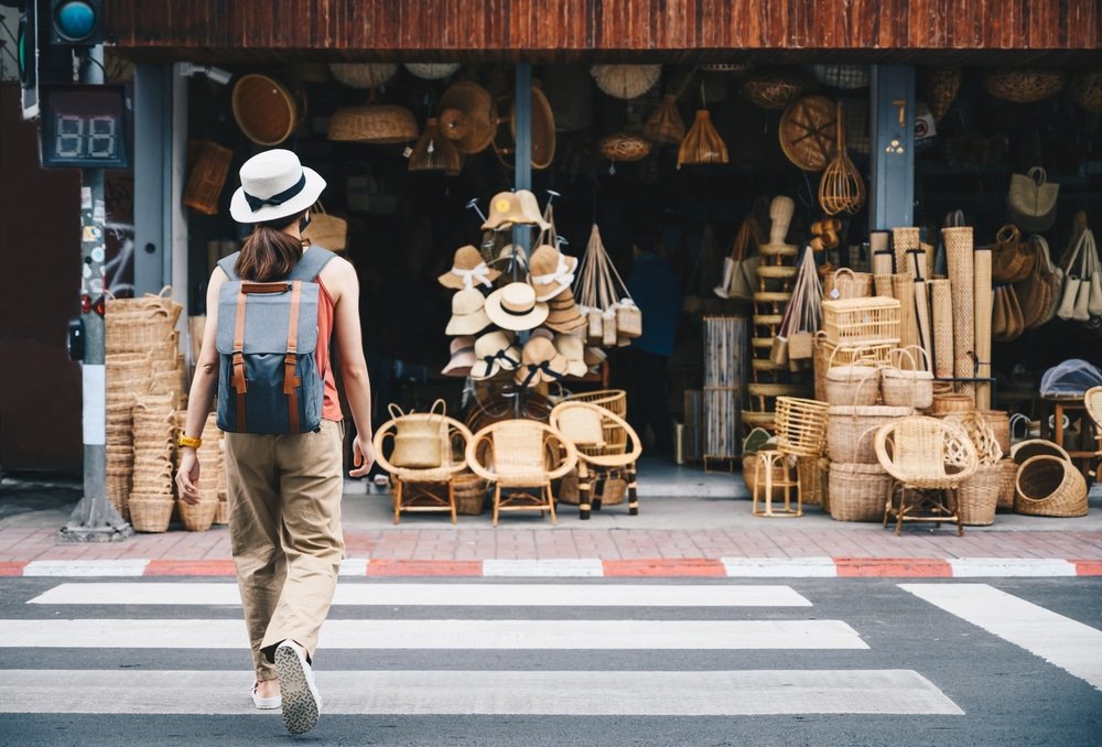 บุคคลที่สวมกระเป๋าเป้และหมวกกำลังข้ามถนนไปยังร้านค้าที่จัดแสดงตะกร้าสานและสินค้าหัตถกรรมอื่น ๆ สะท้อนเสน่ห์ของ ที่เที่ยวเชียงใหม่.