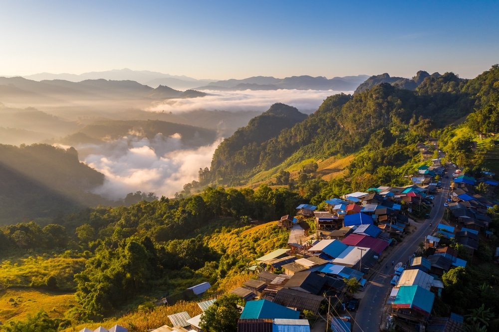 ภาพมุมสูงของหมู่บ้านบนภูเขาที่มีหลังคาหลากสีสัน ล้อมรอบด้วยเนินเขาเขียวขจีและหุบเขาที่เต็มไปด้วยหมอก ภายใต้ท้องฟ้าสีฟ้าใส—ชวนให้นึกถึงเสน่ห์ที่พบได้ในสถานที่ท่องเที่ยวอยุธยาในยามพระอาทิตย์ขึ้น.