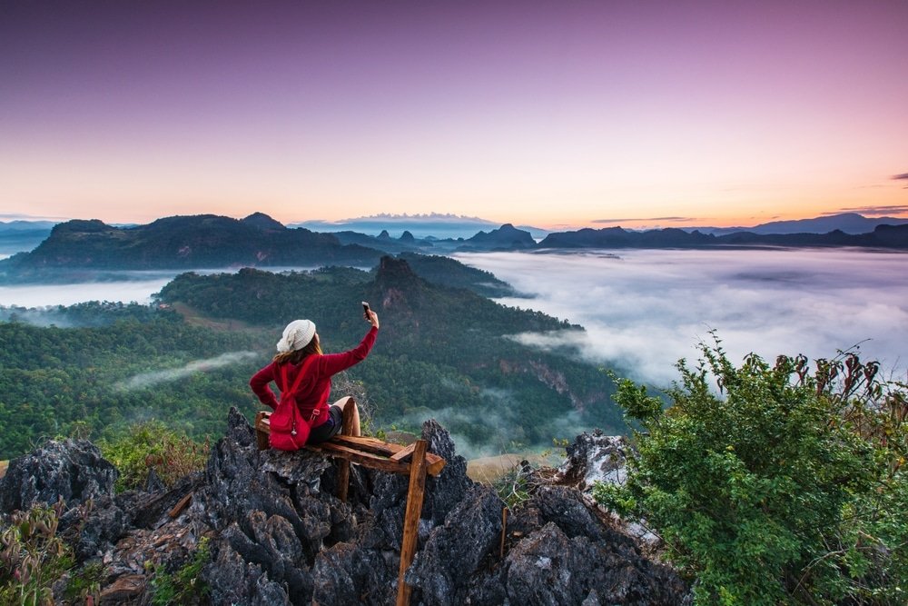 บุคคลในเสื้อแจ็คเก็ตและหมวกสีแดงนั่งอยู่บนพื้นหิน กำลังถ่ายรูปด้วยโทรศัพท์มือถือ มองเห็นหุบเขาและภูเขาที่ปกคลุมด้วยหมอกยามพระอาทิตย์ขึ้น—เป็นภาพที่สร้างแรงบันดาลใจสำหรับผู้ที่วางแผนจะสำรวจ อยุธยาที่เที่ยว ในการผจญภัยครั้งต่อไปของพวกเขา.