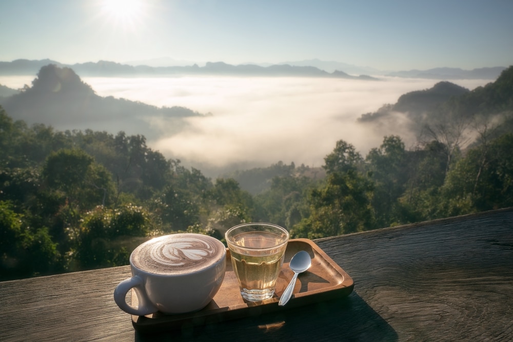 กาแฟหนึ่งถ้วย น้ำหนึ่งแก้ว และช้อนหนึ่งคันบนถาดไม้ รอคุณอยู่ขณะที่คุณชมวิวภูเขาที่ปกคลุมด้วยหมอกยามเช้า—เหมาะสำหรับผู้ที่ต้องการสัมผัส แม่ฮ่องสอนที่เที่ยว พร้อมวิวทิวทัศน์ที่สวยงามตระการตา.