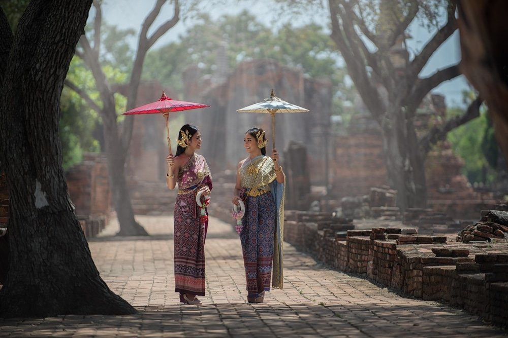 ผู้หญิงสองคนในชุดไทยแบบดั้งเดิมเดินไปด้วยกัน พร้อมถือร่มประดับท่ามกลางซากวัดโบราณและต้นไม้ ณ สถานที่ท่องเที่ยวอยุธยา สะท้อนเสน่ห์อันไร้กาลเวลาของการท่องเที่ยวอยุธยา.