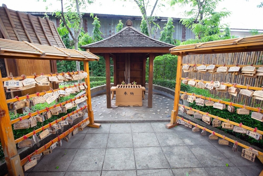 ศาลไม้ขนาดเล็กหลังคาลาดเอียง มีแผ่นป้ายคำอธิษฐานเอมะแขวนเรียงสองแถวด้วยเชือกสีแดง ตั้งอยู่ท่ามกลางสวนที่มีต้นไม้และรั้วไม้ไผ่—สถานที่เงียบสงบเหมาะสำหรับผู้ที่ต้องการ เที่ยวอยุธยา และค้นพบประเพณีอันเป็นเอกลักษณ์.