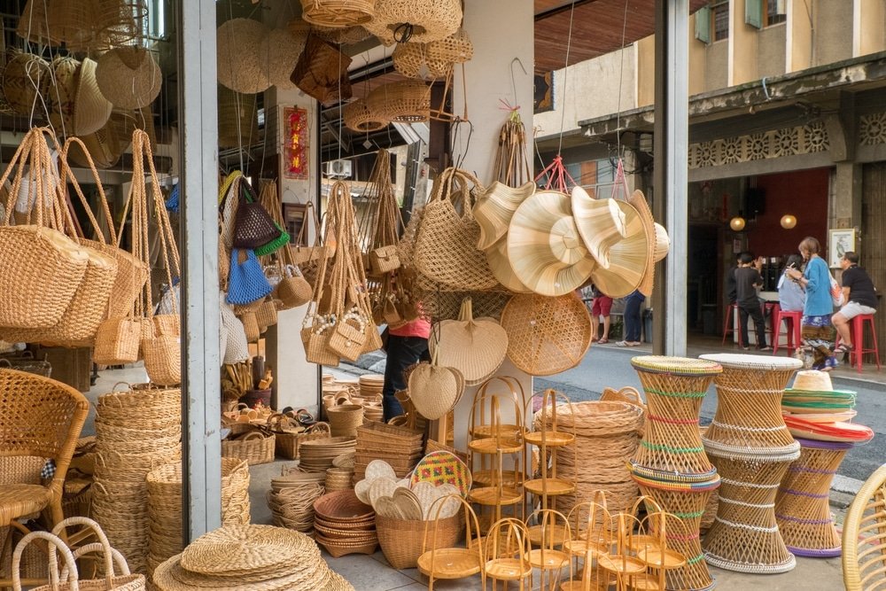 แผงขายของในตลาดถนนแห่งหนึ่งที่สถานที่ท่องเที่ยวเชียงใหม่ แสดงตะกร้า ถุง ถาด และม้านั่งสานหลากหลายชนิดที่ทำจากวัสดุธรรมชาติ โดยมีผู้คนหลายรายอยู่ใกล้เคียง.