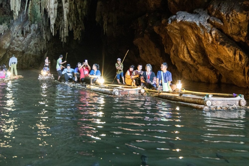 นักท่องเที่ยวนั่งบนแพไม้ไผ่พร้อมกับไกด์ขณะที่พวกเขาเดินทางผ่านถ้ำที่มีแสงสลัวและมีผนังหินและน้ำ เป็นประสบการณ์การท่องเที่ยวอยุธยาที่ไม่เหมือนใครและน่าจดจำ.