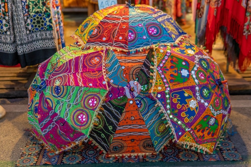 ร่มสามคันที่มีสีสันสดใสและลวดลายซับซ้อนวางอยู่บนพรมลายที่ตลาดกลางแจ้ง—ภาพที่ชวนให้หลงใหลซึ่งคุณอาจพบได้ที่ ที่เที่ยวอยุธยา หนึ่งใน สถานที่ท่องเที่ยวอยุธยา ที่เต็มไปด้วยชีวิตชีวา.