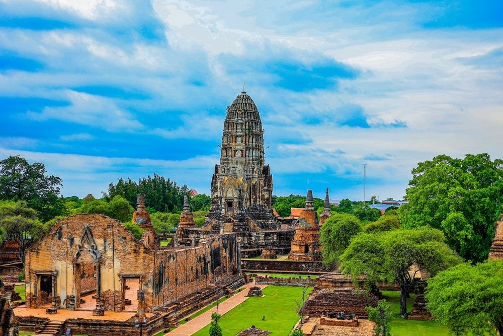 ซากปรักหักพังของวัดโบราณที่มีเจดีย์สูงตรงกลางล้อมรอบด้วยต้นไม้ภายใต้ท้องฟ้าสีฟ้าที่มีเมฆปกคลุม เหมาะสำหรับผู้ที่มองหาสถานที่ท่องเที่ยวในอยุธยาหรือวางแผนที่จะเที่ยวอยุธยาท่ามกลางความงามทางประวัติศาสตร์.