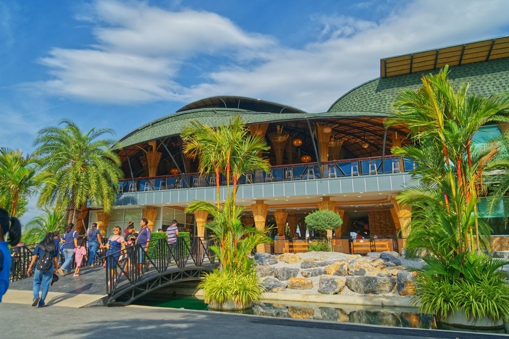 ผู้คนเดินบนสะพานมุ่งหน้าไปยังอาคารขนาดใหญ่ทันสมัยที่มีหลังคาโดมสีเขียว ล้อมรอบด้วยต้นปาล์มและหินที่จัดแต่งภูมิทัศน์ไว้อย่างสวยงามใต้ท้องฟ้าสีคราม—เป็นหนึ่งในสถานที่ท่องเที่ยวอันเป็นเอกลักษณ์ของอยุธยาสำหรับผู้ที่ต้องการเที่ยวชมเมืองเก่าอยุธยา.