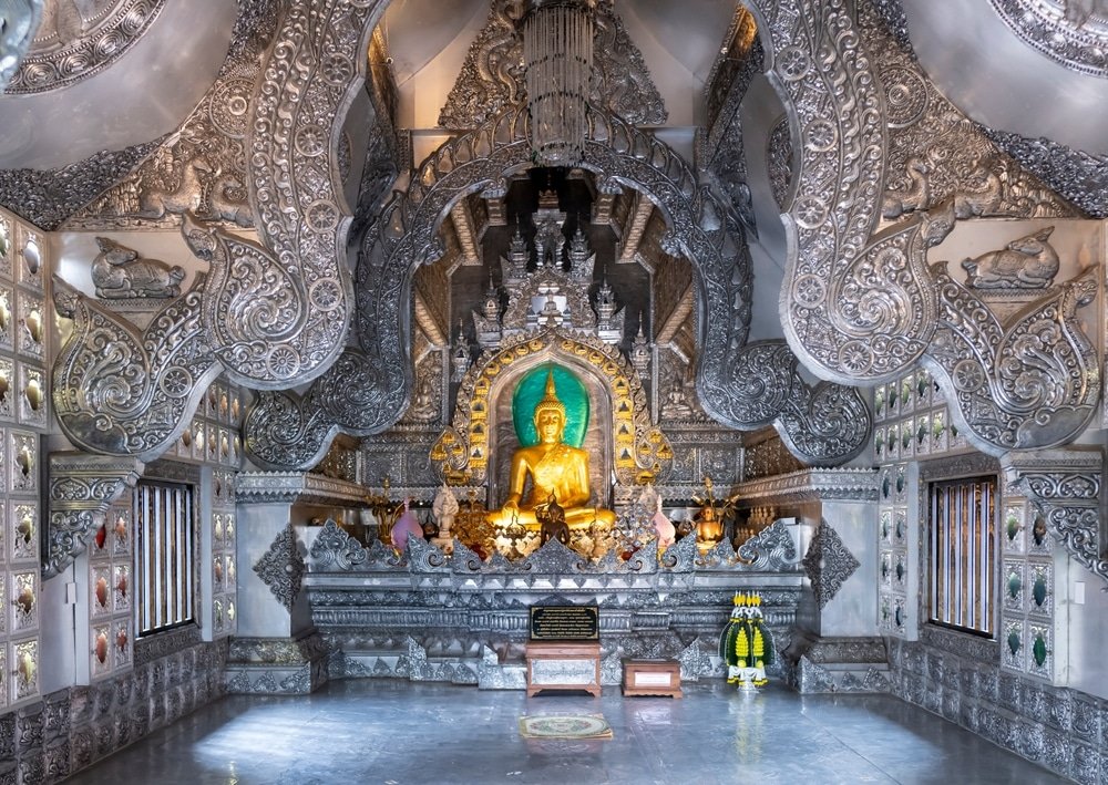 ภายในวัดเงินอันวิจิตรตระการตาในเชียงใหม่ สถานที่ท่องเที่ยวที่มีชื่อเสียง ประดับด้วยงานแกะสลักที่ประณีตละเอียดอ่อน โดดเด่นด้วยพระพุทธรูปทองคำตั้งอยู่ตรงกลางแท่นบูชา ล้อมรอบด้วยองค์ประกอบตกแต่งและเครื่องบูชา.