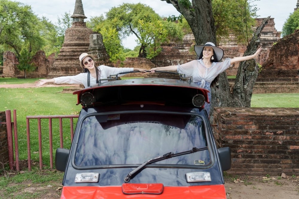 ผู้หญิงสองคนสวมหมวกกันแดดยืนอยู่ข้างรถตุ๊กตุ๊กสีแดง ยิ้มและโพสท่าด้วยแขนเหยียดออกท่ามกลางซากปรักหักพังโบราณและพืชพรรณเขียวขจี—ถ่ายทอดจิตวิญญาณแห่งความสนุกสนานของการเที่ยวอยุธยา ณ หนึ่งในสถานที่ท่องเที่ยวอยุธยาที่ต้องไปเยือน.