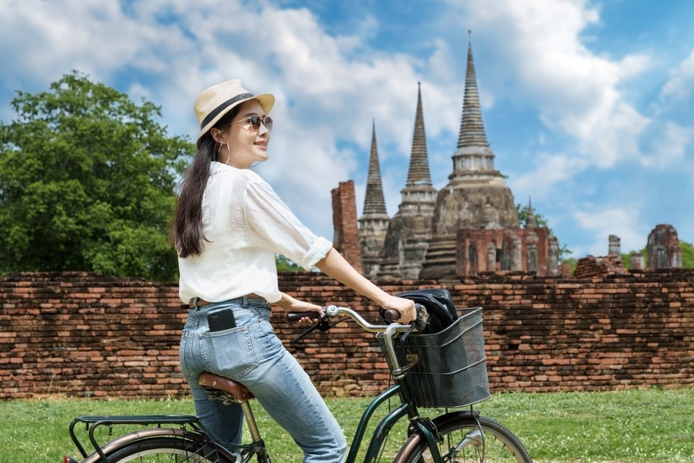 ผู้หญิงสวมหมวกและแว่นกันแดดขี่จักรยานผ่านซากอิฐโบราณและยอดเจดีย์แหลมภายใต้ท้องฟ้าที่มีเมฆบางส่วน จับภาพเสน่ห์ของการเที่ยวอยุธยา.