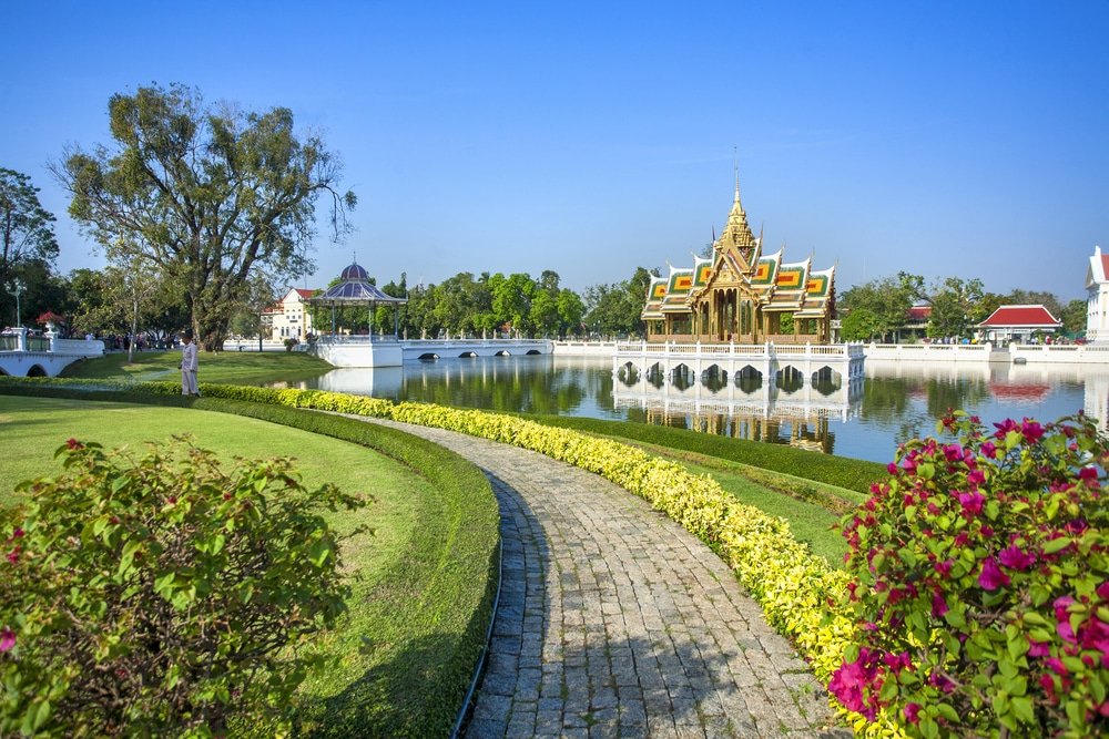 สวนที่จัดแต่งอย่างสวยงามพร้อมทางเดินหินโค้งไปยังศาลาไทยแบบดั้งเดิมบนสระน้ำ—หนึ่งในสถานที่ท่องเที่ยวที่น่าหลงใหลในอยุธยา—ล้อมรอบด้วยพุ่มไม้ตัดแต่งอย่างประณีตและดอกไม้หลากสีภายใต้ท้องฟ้าสีครามสดใส.