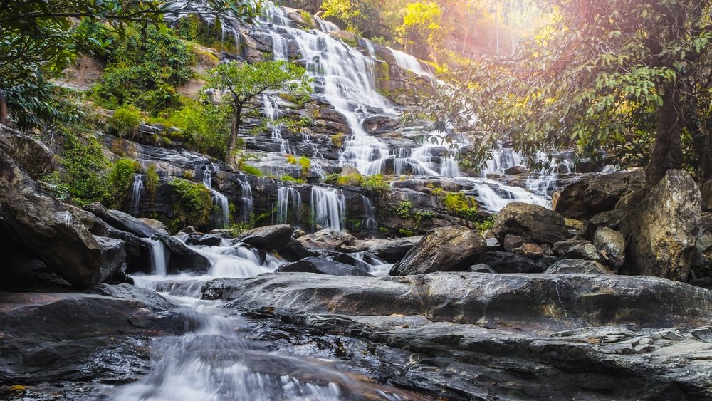 น้ำตกหลายชั้นไหลลดหลั่นกันลงสู่โขดหิน ท่ามกลางพืชพรรณเขียวชอุ่ม ส่องประกายแสงแดดที่ลอดผ่านยอดไม้ด้านบน—สถานที่ลับที่เหมาะสำหรับผู้ที่ต้องการสำรวจแหล่งท่องเที่ยวเชียงใหม่และสัมผัสความงามตามธรรมชาติของเชียงใหม่.