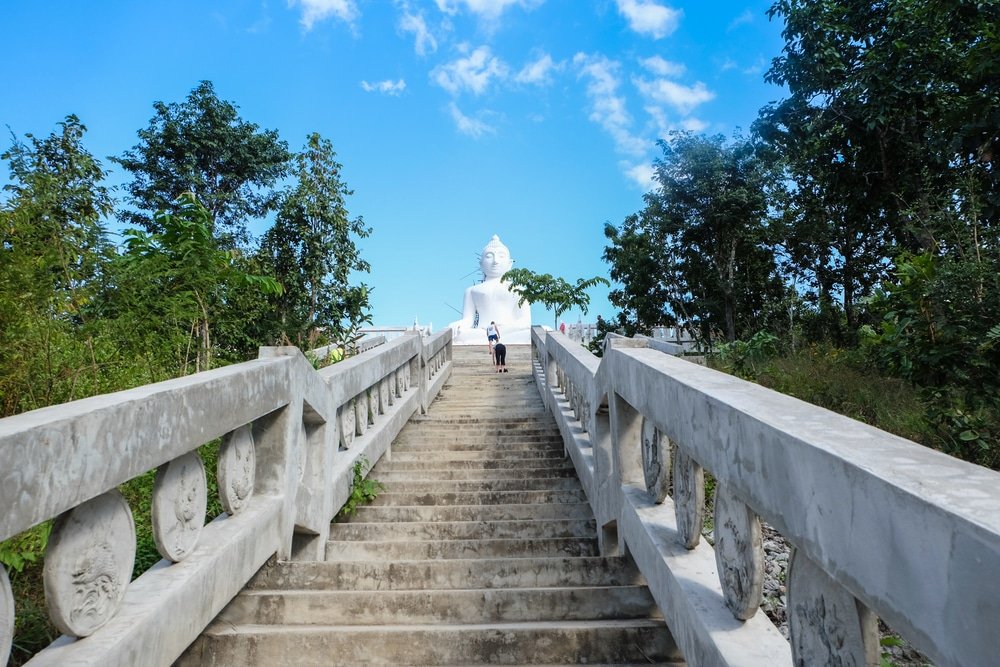 บันไดคอนกรีตกว้างทอดยาวขึ้นเนินไปยังพระพุทธรูปขนาดใหญ่สีขาว ล้อมรอบด้วยต้นไม้ใต้ท้องฟ้าสีฟ้าสดใสที่มีเมฆกระจายอยู่—สถานที่ท่องเที่ยวอยุธยาที่เป็นสัญลักษณ์ เหมาะสำหรับผู้ที่วางแผนจะเที่ยวอยุธยา.