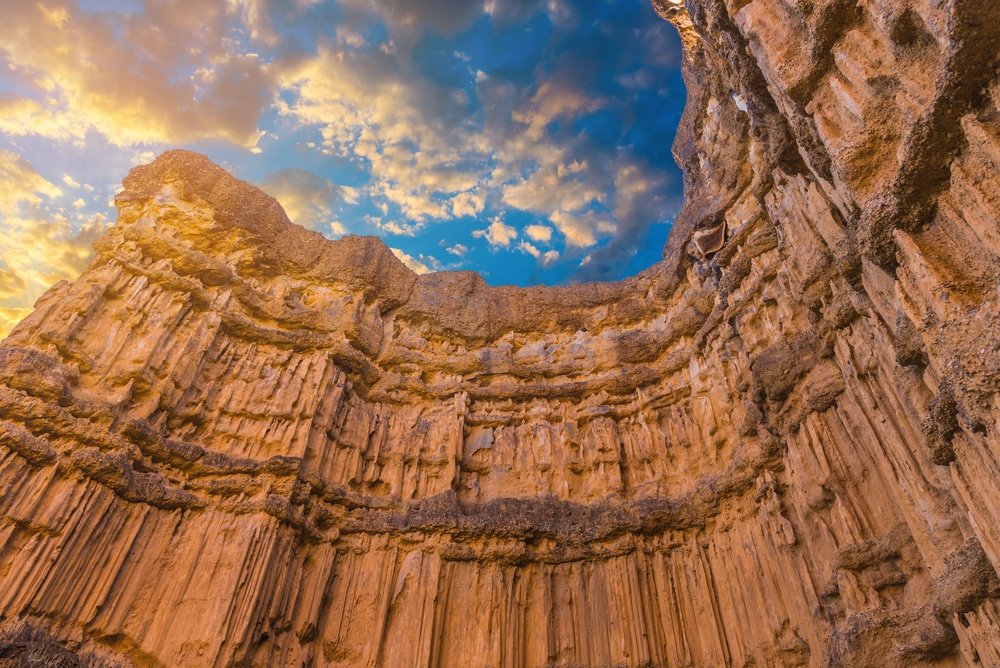 หน้าผาหินสูงชันซ้อนชั้นกันทอดตัวขึ้นสู่ท้องฟ้าที่มีเมฆบางส่วนปกคลุมยามพระอาทิตย์ตกดิน ณ หนึ่งในสถานที่ท่องเที่ยวเชียงใหม่ แสดงให้เห็นลายเส้นสีส้มและสีเบจตัดกับพื้นผิวขรุขระอย่างงดงาม.