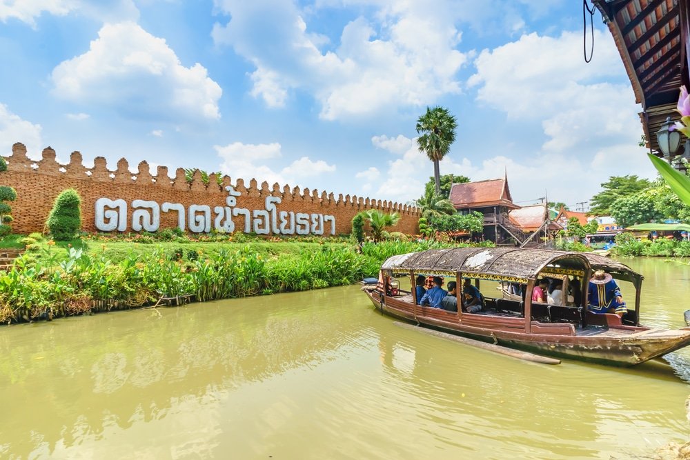 เรือไม้ลำหนึ่งพร้อมผู้โดยสารแล่นไปตามคลองข้างสวนที่เขียวชอุ่มและกำแพงอิฐขนาดใหญ่ที่มีอักษรไทย ภายใต้ท้องฟ้าที่มีเมฆบางส่วน—ภาพที่น่ารักเหมาะสำหรับผู้ที่ต้องการเที่ยวอยุธยา.