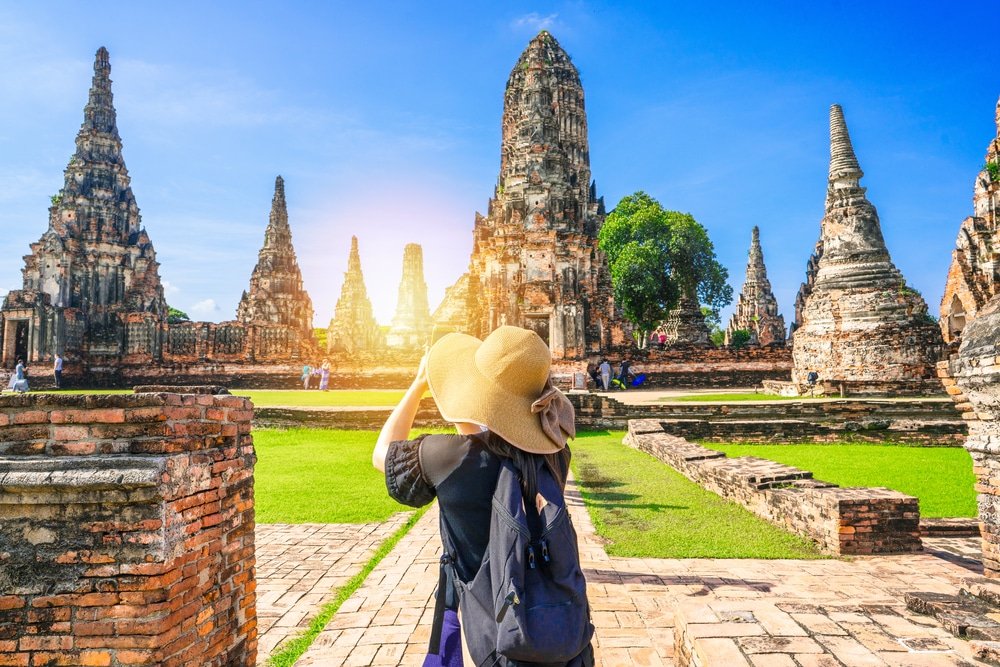 บุคคลหนึ่งสวมหมวกและสะพายเป้ยืนอยู่หน้าซากปรักหักพังของวัดโบราณภายใต้ท้องฟ้าสีฟ้าใส พร้อมที่จะสำรวจสถานที่ท่องเที่ยวอยุธยาด้วยแสงแดดที่ส่องสว่าง.