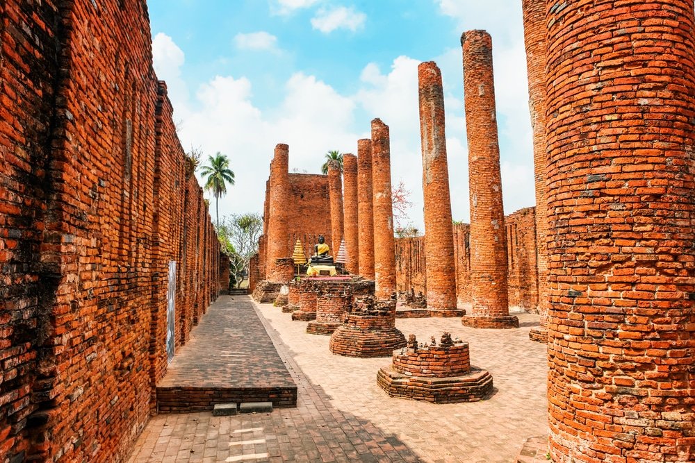 เสาอิฐโบราณและซากปรักหักพังพร้อมพระพุทธรูปอยู่ตรงกลางภายใต้ท้องฟ้าสีคราม ถ่ายทอดความงามอันไร้กาลเวลาของสถานที่ท่องเที่ยวอยุธยา ณ วัดประวัติศาสตร์อันน่าทึ่งแห่งนี้.