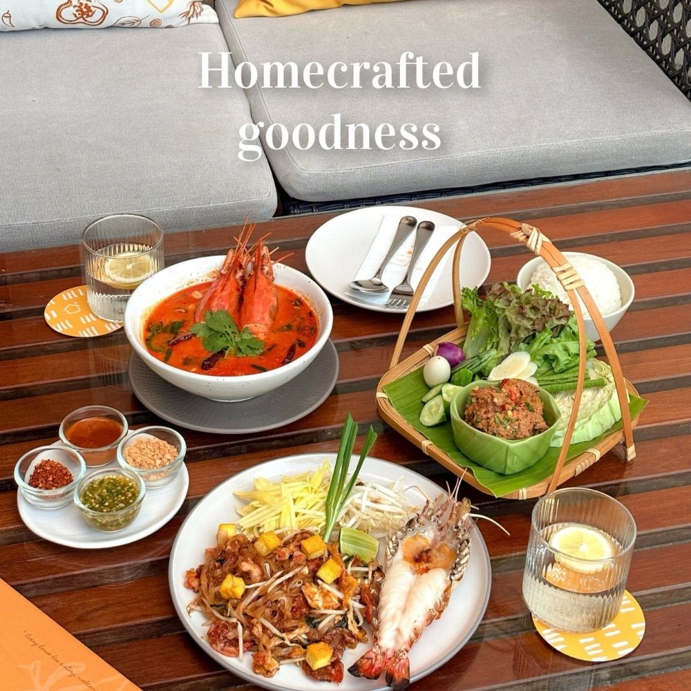 โต๊ะไม้ที่จัดวางอาหารไทย—ซุปกุ้ง, ผัดไทยกุ้ง, จานผักรวม, ซอส, และเครื่องดื่ม—สะท้อนถึง "ความอร่อยแบบโฮมเมด" ที่สมบูรณ์แบบสำหรับการเติมพลังให้กับการผจญภัยครั้งต่อไปของคุณที่ เที่ยวอยุธยา.