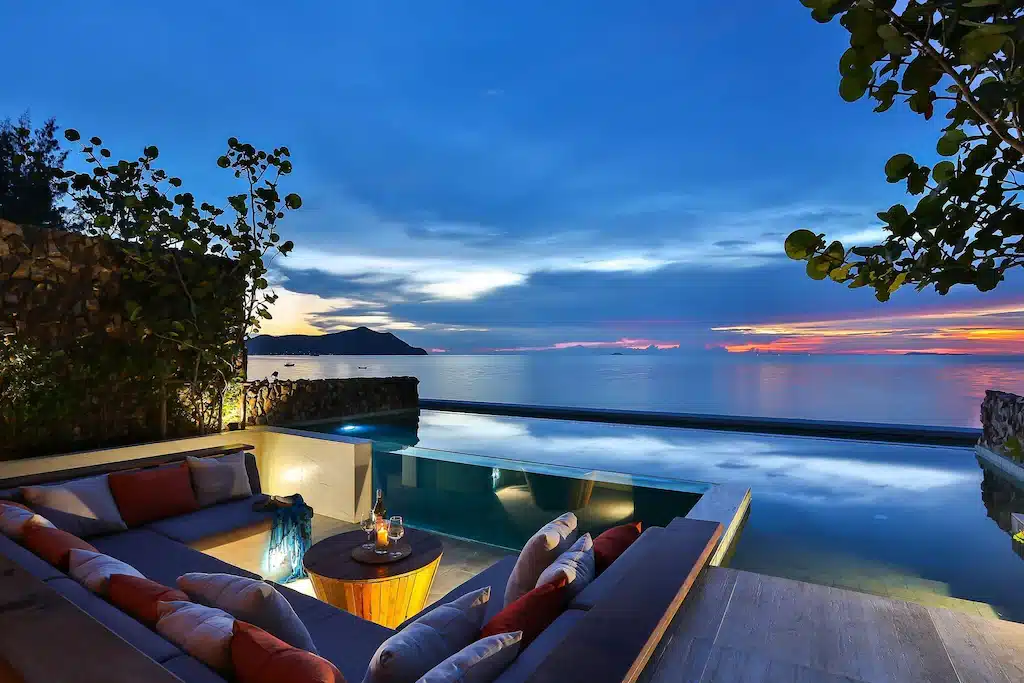 พื้นที่นั่งเล่นกลางแจ้งของโรงแรมติดทะเลพัทยาแห่งนี้ ตั้งอยู่ริมสระว่ายน้ำแบบอินฟินิตี้ มองเห็นวิวทะเลที่สงบในยามพระอาทิตย์ตก พร้อมเครื่องดื่มบนโต๊ะและต้นไม้ที่ล้อมรอบทัศนียภาพอันเงียบสงบ.