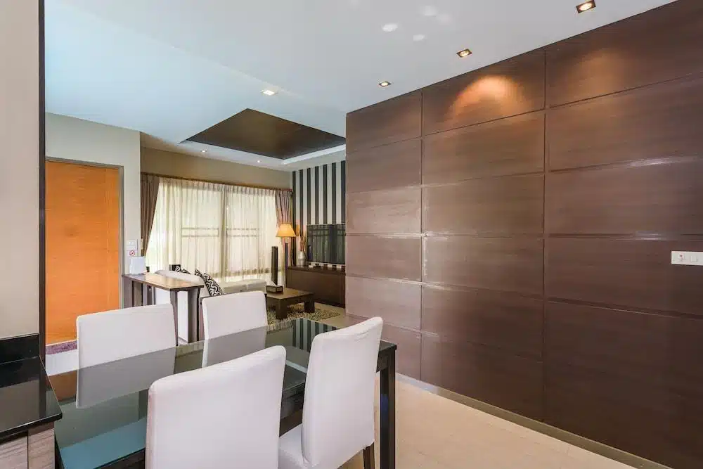 พื้นที่รับประทานอาหารสมัยใหม่พร้อมโต๊ะกระจกและเก้าอี้สีขาวหกตัว ติดกับพื้นที่นั่งเล่น หน้าต่างบานใหญ่พร้อมผ้าม่าน และผนังไม้ตกแต่ง—เหมาะสำหรับการพักผ่อนหลังจากสำรวจโรงแรมติดทะเลพัทยาหรือที่พักพัทยาติดทะเล.