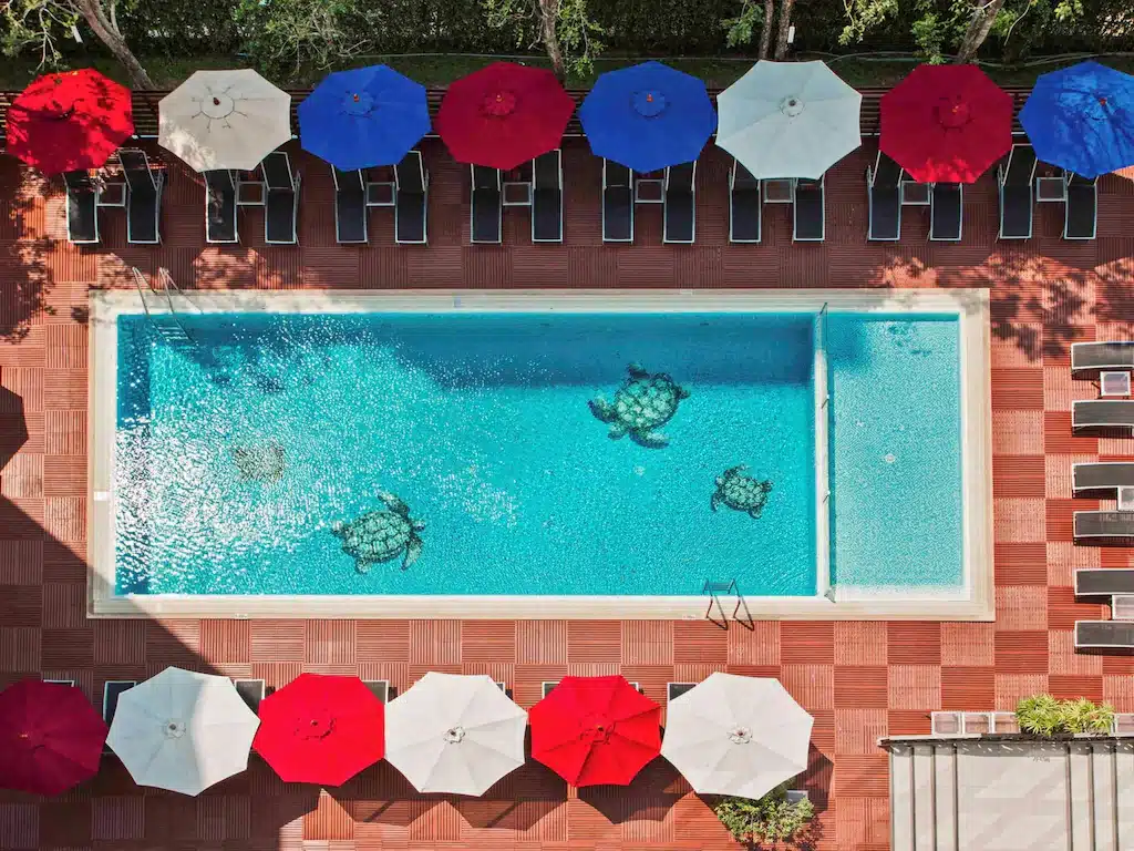 สระว่ายน้ำกลางแจ้งรูปสี่เหลี่ยมผืนผ้าที่ที่พักพัทยาติดทะเลรายล้อมไปด้วยร่มสีแดง ขาว และน้ำเงิน พร้อมเก้าอี้อาบแดด มองจากมุมสูง.