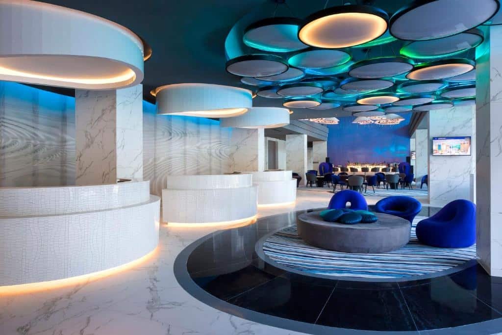 ล็อบบี้โรงแรมสมัยใหม่พร้อมเคาน์เตอร์ต้อนรับสีขาวรูปวงกลม, ไฟเน้นสีน้ำเงิน, พื้นหินอ่อน, และที่นั่งร่วมสมัยรวมถึงเก้าอี้สีน้ำเงินและโซฟาทรงกลมที่โรงแรมติดทะเลพัทยาเพื่อการพักผ่อนริมทะเลอย่างมีสไตล์.