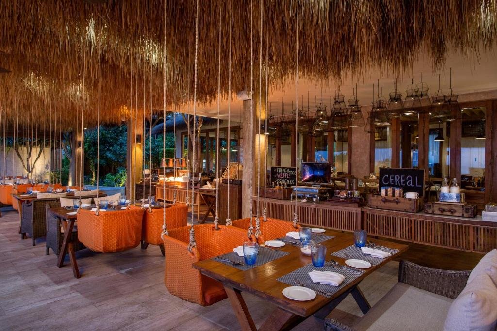 ร้านอาหารธีมเขตร้อนในโรงแรมติดทะเลพัทยา มีเก้าอี้สีส้ม โต๊ะไม้ โคมไฟแขวน เพดานจากหญ้าคา และพื้นที่บุฟเฟ่ต์อาหารเช้าที่มีป้ายว่า "ซีเรียล" และ "โยเกิร์ตสด.