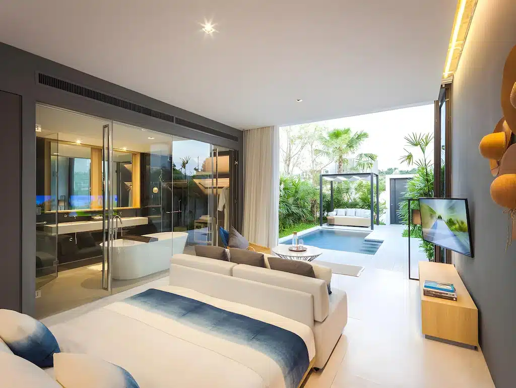 ห้องพักโรงแรมสมัยใหม่ที่ โรงแรมพัทยาติดทะเล พร้อมห้องน้ำผนังกระจก เตียงนอนสบาย พื้นที่นั่งเล่น โทรทัศน์ และประตูบานเลื่อนสู่ระเบียงส่วนตัวพร้อมสระว่ายน้ำและที่นั่งกลางแจ้ง—ทั้งหมดล้อมรอบด้วยพืชพรรณเขียวขจี.
