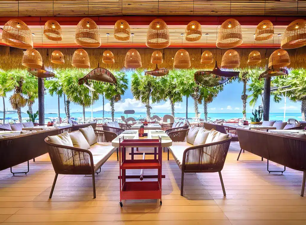 พื้นที่นั่งรับประทานอาหารกลางแจ้งพร้อมเก้าอี้หวาย ม้านั่งบุเบาะ และโคมไฟแขวน ล้อมรอบด้วยต้นปาล์มและมองเห็นวิวทะเล เหมาะอย่างยิ่งสำหรับการพักผ่อนที่ ที่พักพัทยาติดทะเล ท่ามกลางท้องฟ้าสีครามสดใส.