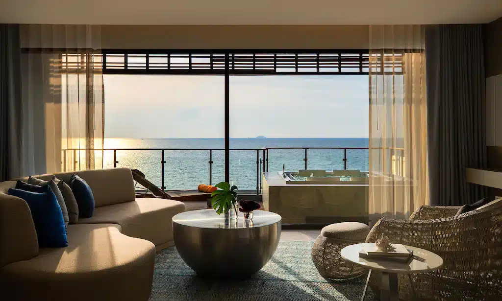 ห้องนั่งเล่นสไตล์โมเดิร์นพร้อมหน้าต่างบานใหญ่ที่มองเห็นระเบียง, อ่างน้ำร้อนกลางแจ้ง, และวิวทะเลยามพระอาทิตย์ตก เหมาะสำหรับผู้ที่ต้องการโรงแรมติดทะเลพัทยาพร้อมเฟอร์นิเจอร์สมัยใหม่และโทนสีกลางที่ผ่อนคลายทั่วทั้งพื้นที่.