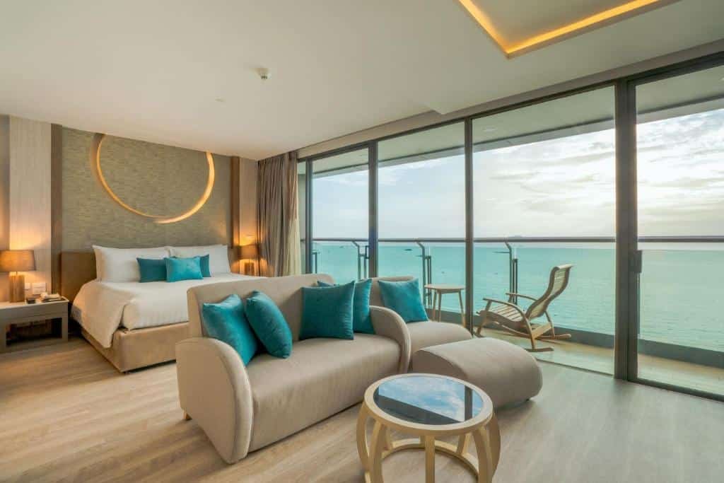 ห้องพักโรงแรมสมัยใหม่พร้อมเตียงขนาดใหญ่ โซฟา และผนังกระจกที่เปิดออกสู่ระเบียงมองเห็นวิวทะเล เหมาะสำหรับผู้ที่ต้องการที่พักพัทยาติดทะเล การตกแต่งเป็นโทนสีกลางๆ พร้อมด้วยสีเขียวอมฟ้าและพื้นไม้.