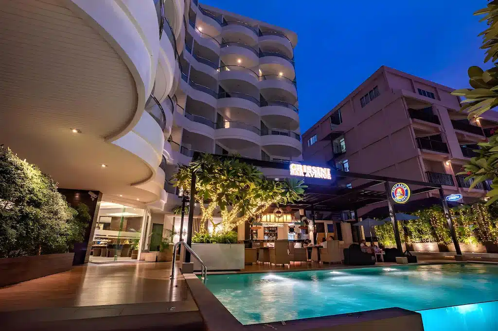 บริเวณสระว่ายน้ำกลางแจ้งของโรงแรมพัทยาติดทะเลสไตล์โมเดิร์นในช่วงพลบค่ำ พร้อมร้านอาหารและบาร์ที่เปิดไฟสว่าง รายล้อมด้วยอาคารสูงหลายชั้นและภูมิทัศน์ที่ประดับไฟสวยงาม.