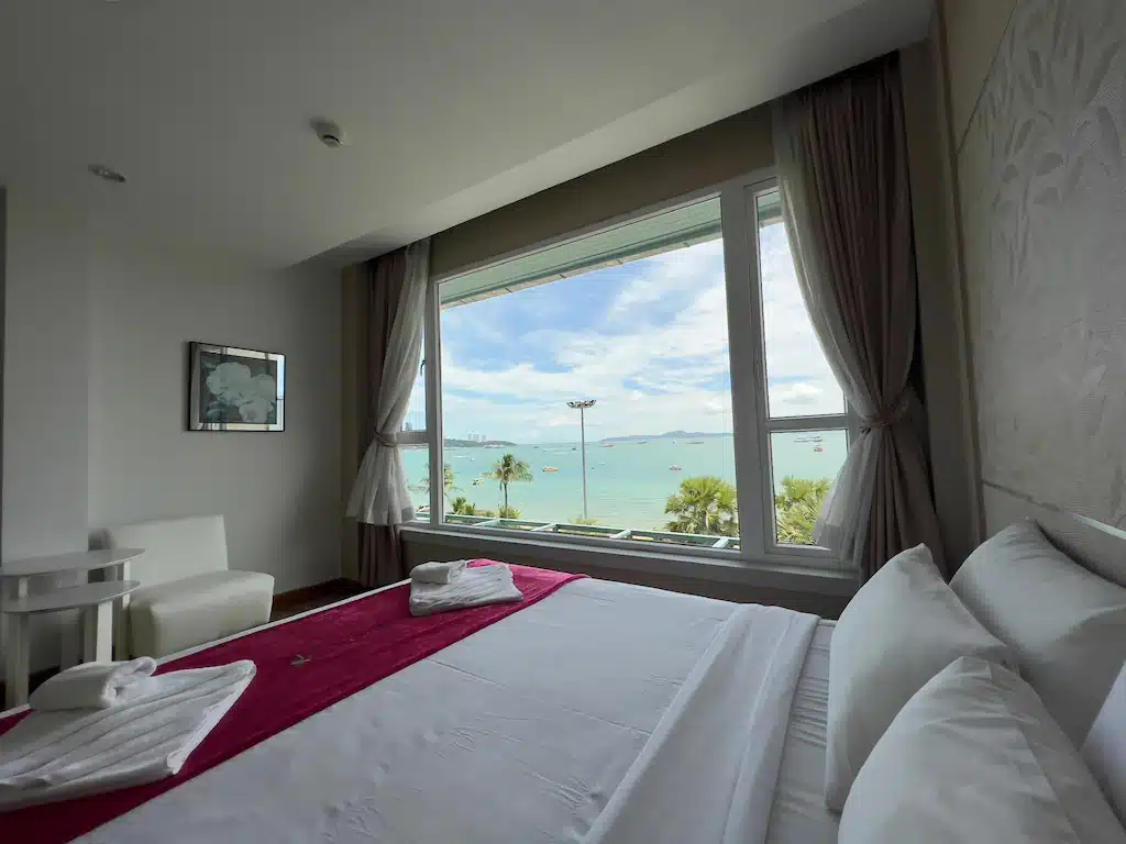 ห้องพักในโรงแรมที่มีเตียงปูเรียบร้อย หน้าต่างบานใหญ่เปิดรับวิวทะเล ต้นปาล์ม และท้องฟ้าสีครามด้านนอก—เหมาะอย่างยิ่งสำหรับผู้ที่กำลังมองหา โรงแรมพัทยาติดทะเล สำหรับการพักผ่อนริมทะเลอย่างแท้จริง.