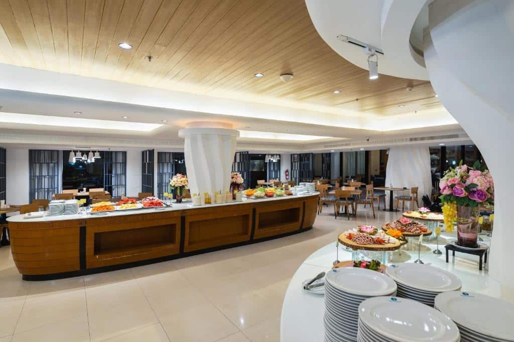 ห้องอาหารบุฟเฟ่ต์กว้างขวางในบรรยากาศทันสมัยและสว่างไสว ตกแต่งด้วยไม้เป็นส่วนประกอบ—เหมาะสำหรับแขกจากที่พักพัทยาติดทะเล มีเคาน์เตอร์ยาวที่จัดเรียงอาหารหลากหลายชนิด ดอกไม้สด และจานซ้อนเรียงไว้อย่างเรียบร้อย.