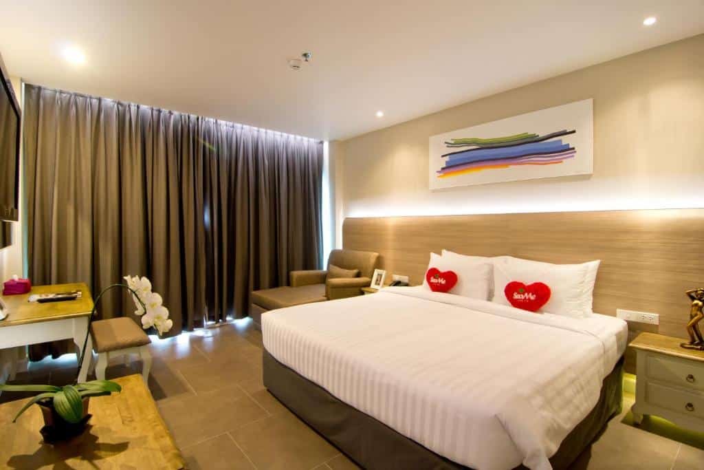 ห้องพักโรงแรมสมัยใหม่พร้อมเตียงขนาดใหญ่ หมอนรูปหัวใจสีแดงสองใบ เก้าอี้นวม โต๊ะทำงาน ต้นกล้วยไม้ และศิลปะนามธรรมบนผนัง เหมาะสำหรับผู้ที่มองหา โรงแรมพัทยาติดทะเล ที่มีสไตล์และความสะดวกสบาย ม่านสีเข้มปิดหน้าต่าง.