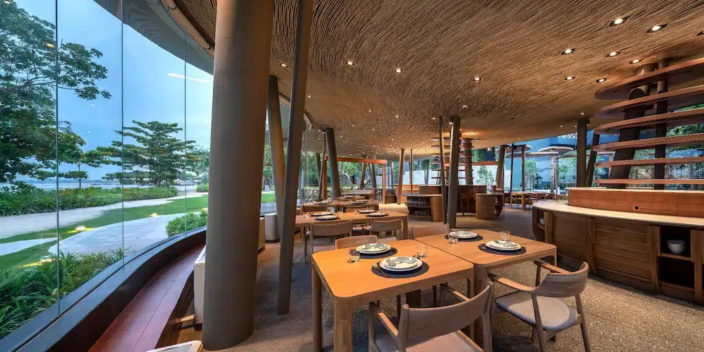 ร้านอาหารสมัยใหม่พร้อมโต๊ะและเก้าอี้ไม้ ตกแต่งด้วยจานและช้อนส้อม มีหน้าต่างบานใหญ่จากพื้นจรดเพดานที่มองเห็นสวนและพื้นที่ชายหาด ณ ที่พักติดทะเลพัทยาที่มีสไตล์.