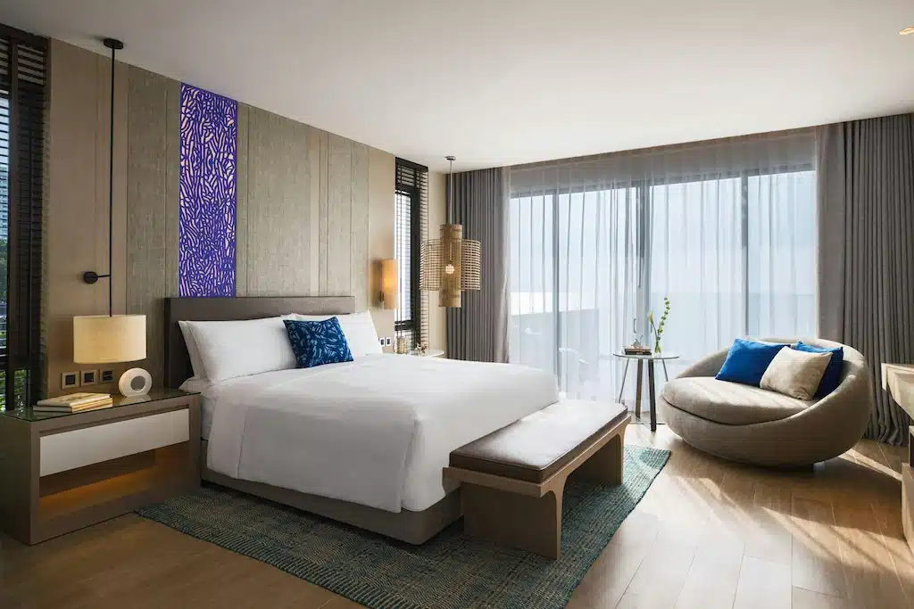 ห้องพักโรงแรมสมัยใหม่พร้อมเตียงขนาดใหญ่, รายละเอียดสีน้ำเงิน, เก้าอี้ทรงกลม, เฟอร์นิเจอร์ไม้, และหน้าต่างบานใหญ่จากพื้นจรดเพดานพร้อมผ้าม่านบาง—เหมาะสำหรับการเข้าพักของคุณที่ โรงแรมพัทยาติดทะเล.