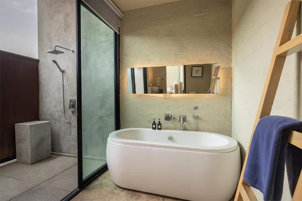 ห้องน้ำสไตล์โมเดิร์นในโรงแรมติดทะเลพัทยา พร้อมอ่างอาบน้ำทรงรีแบบลอยตัว ก๊อกน้ำติดผนัง กระจกบานใหญ่ ผนังหินเปลือย และพื้นที่อาบน้ำกระจกใส ผ้าเช็ดตัวแขวนบนบันไดไม้ เพิ่มเสน่ห์แบบชายทะเล.