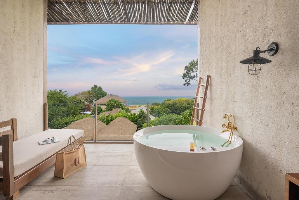 อ่างอาบน้ำทรงกลมบนระเบียงพร้อมวิวทะเลและท้องฟ้า ณ โรงแรมติดทะเลพัทยา ข้างม้านั่งไม้ บันได โคมไฟติดผนัง และกระเป๋าถือสีเบจ.